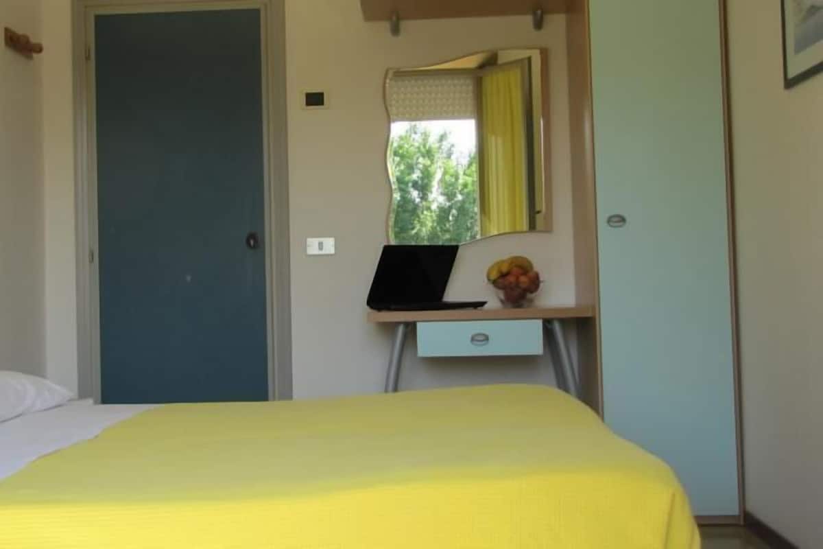A bedroom with a bed, a desk with a laptop, a mirror, and a door.