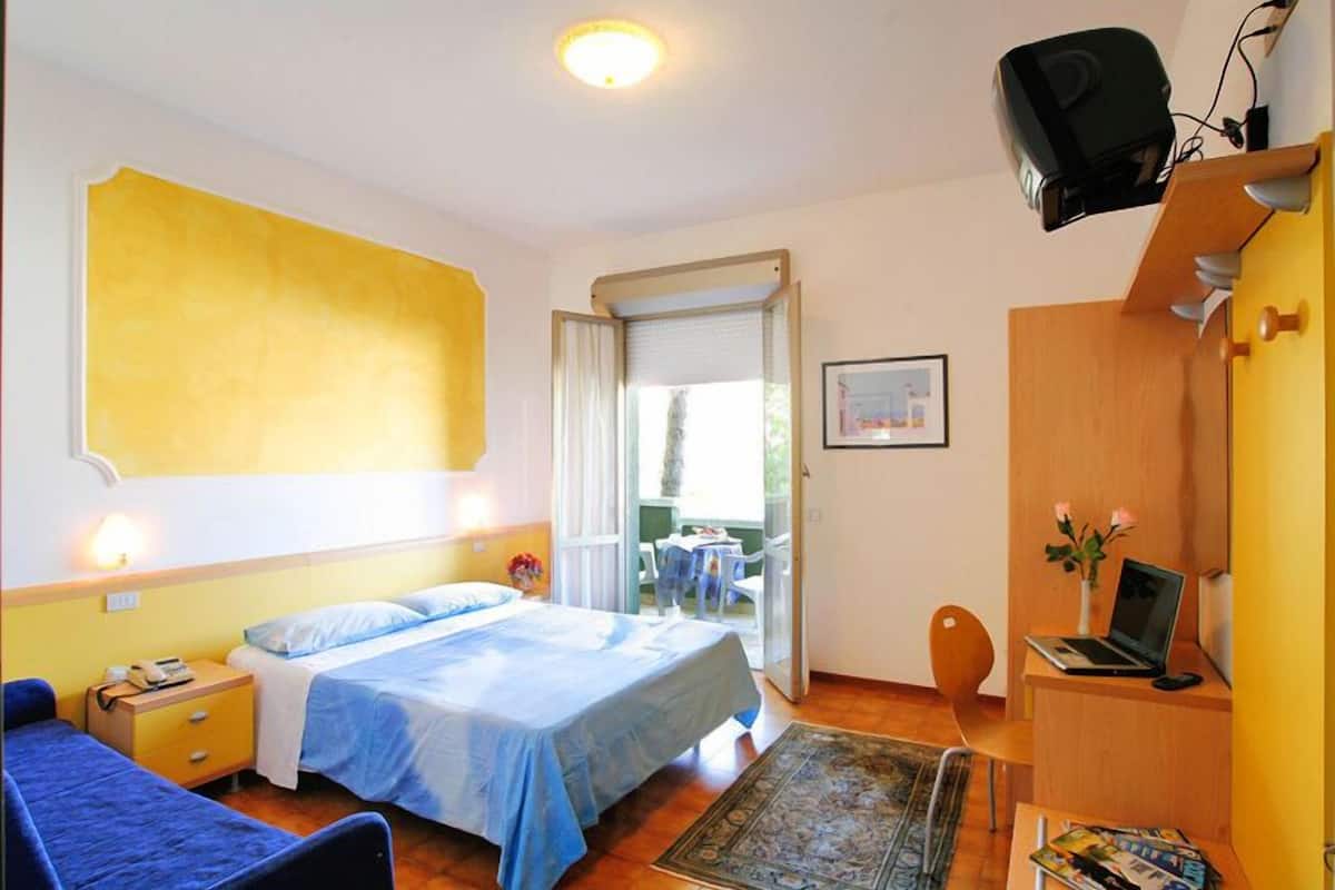 A hotel room with a bed, a desk with a laptop, a chair, a small table with flowers, and a window with curtains.