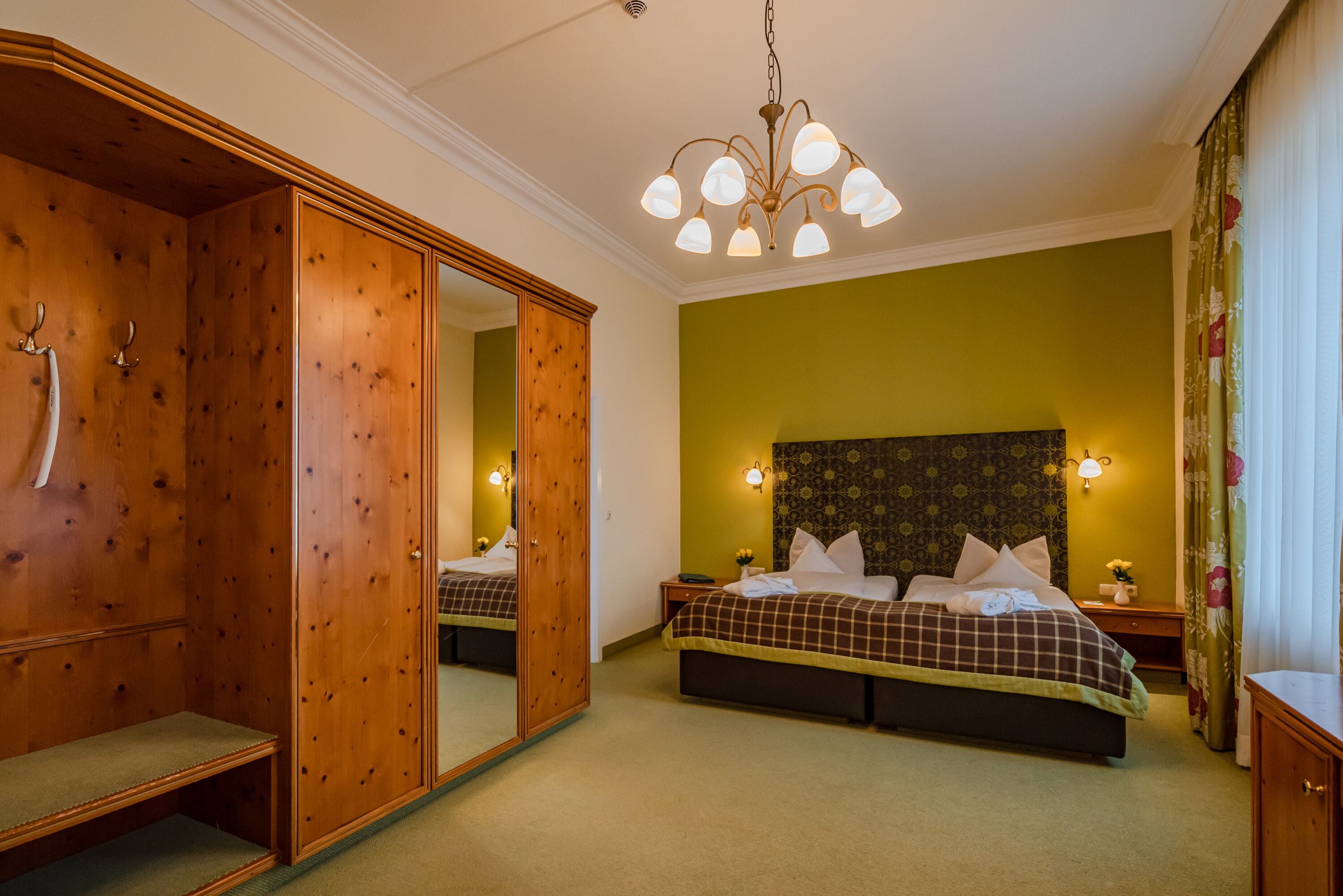 Une chambre avec une grande armoire, un lit recouvert d’une courtepointe à carreaux et un lustre.