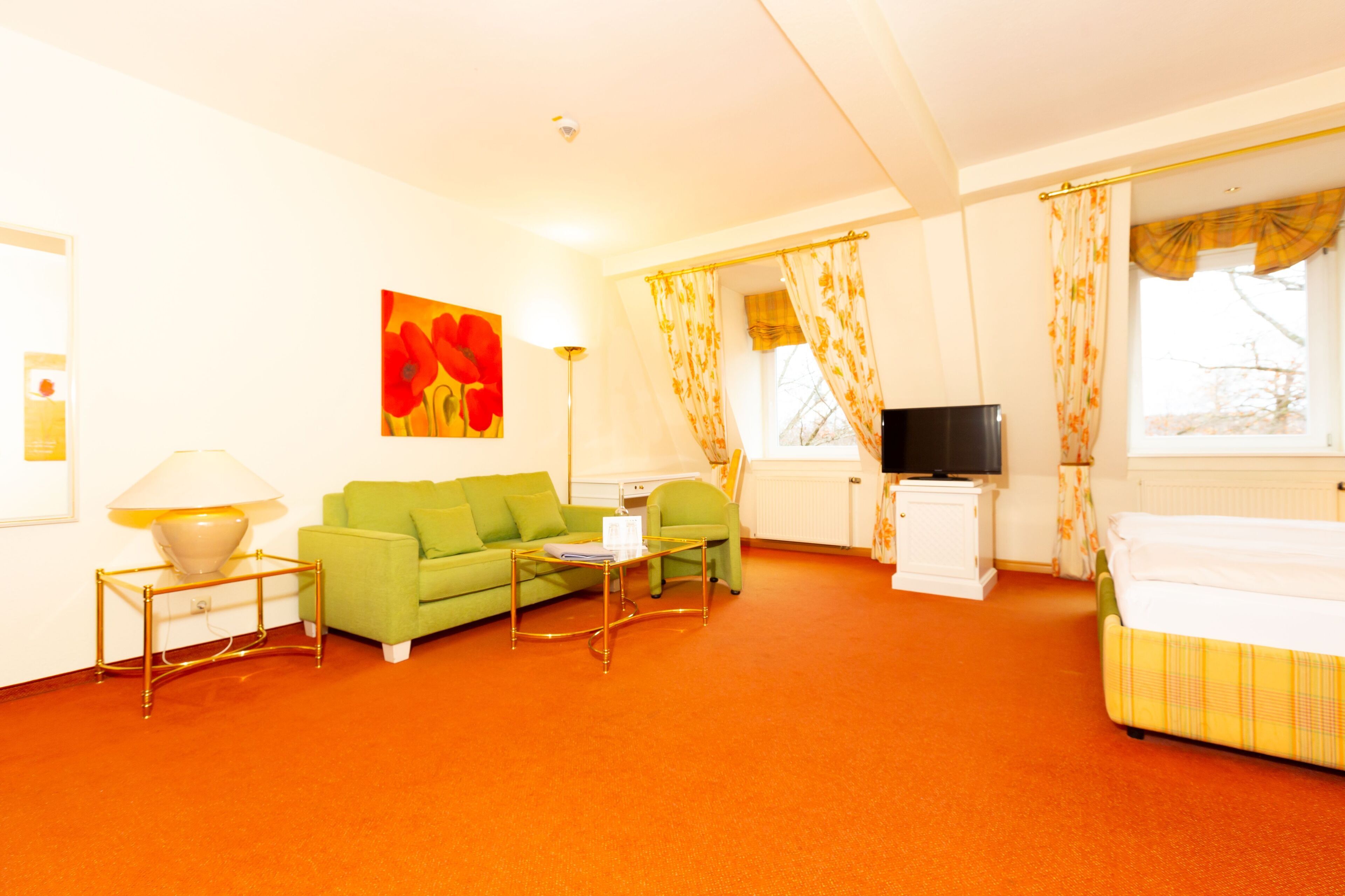 Habitación de hotel con un sofá verde, una mesita, una lámpara, un televisor, una cama y una ventana con cortinas.