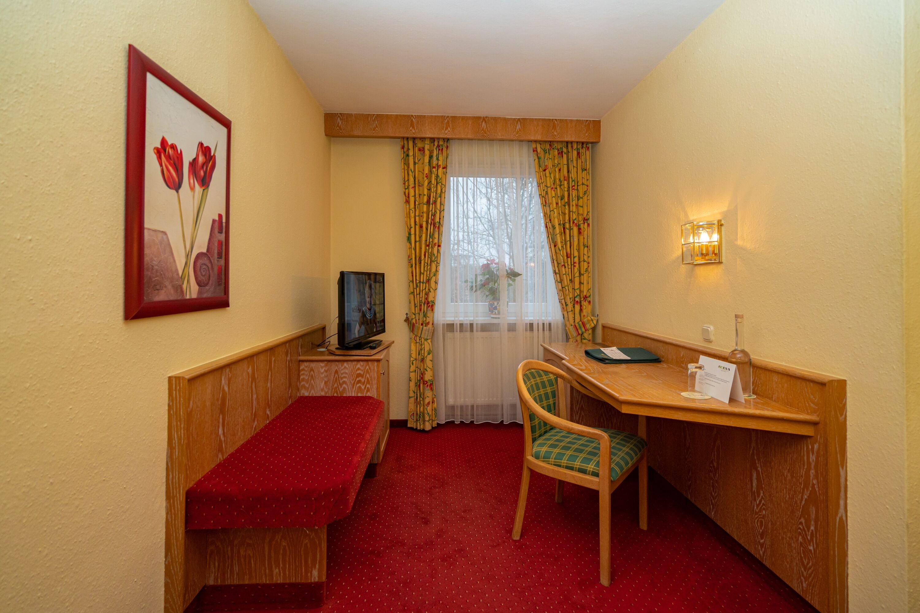 A small room with a wooden desk, a chair, a bench, a television, and a painting on the wall.
