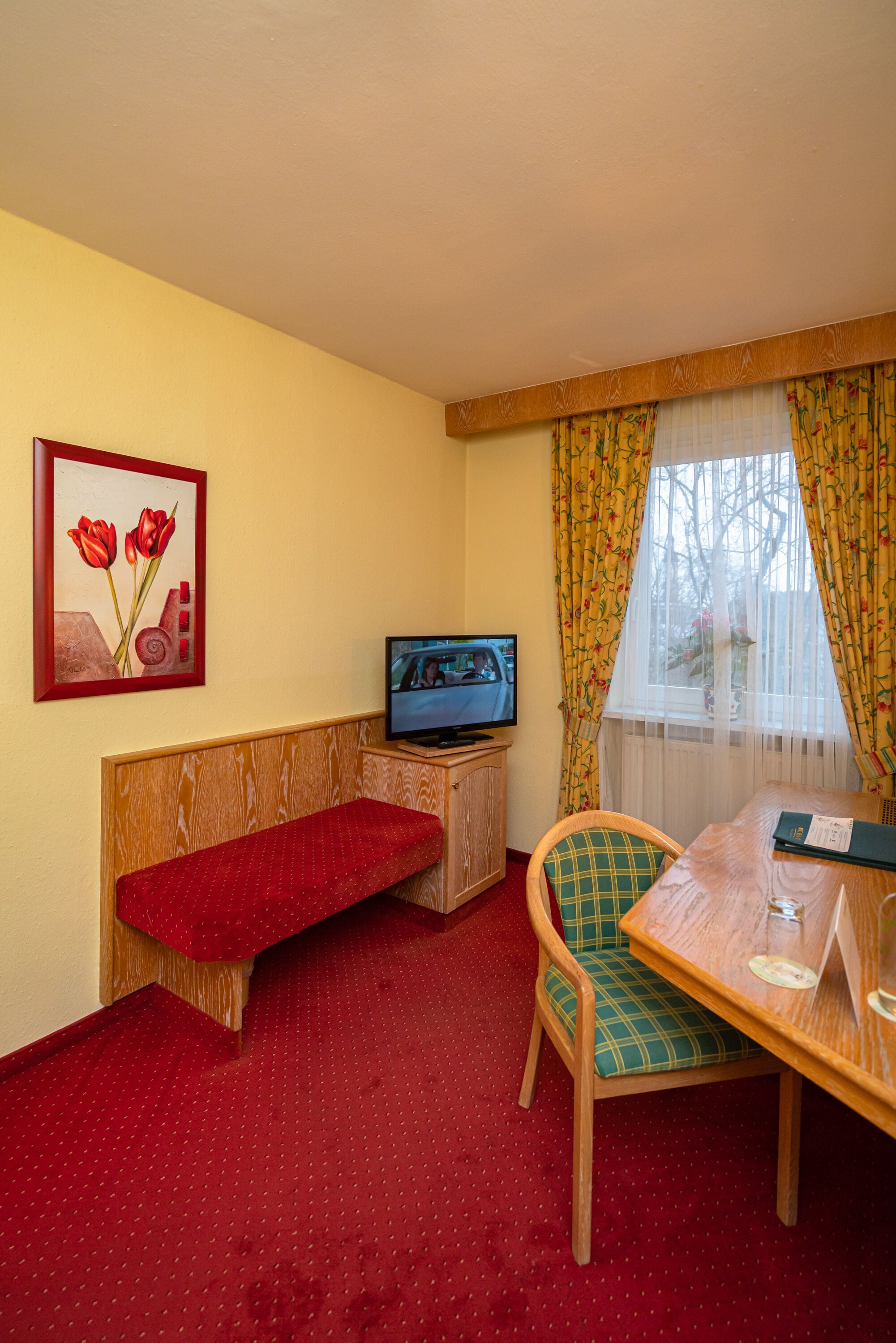 A small room with a wooden bench, a table with a chair, a television, and a window with curtains.