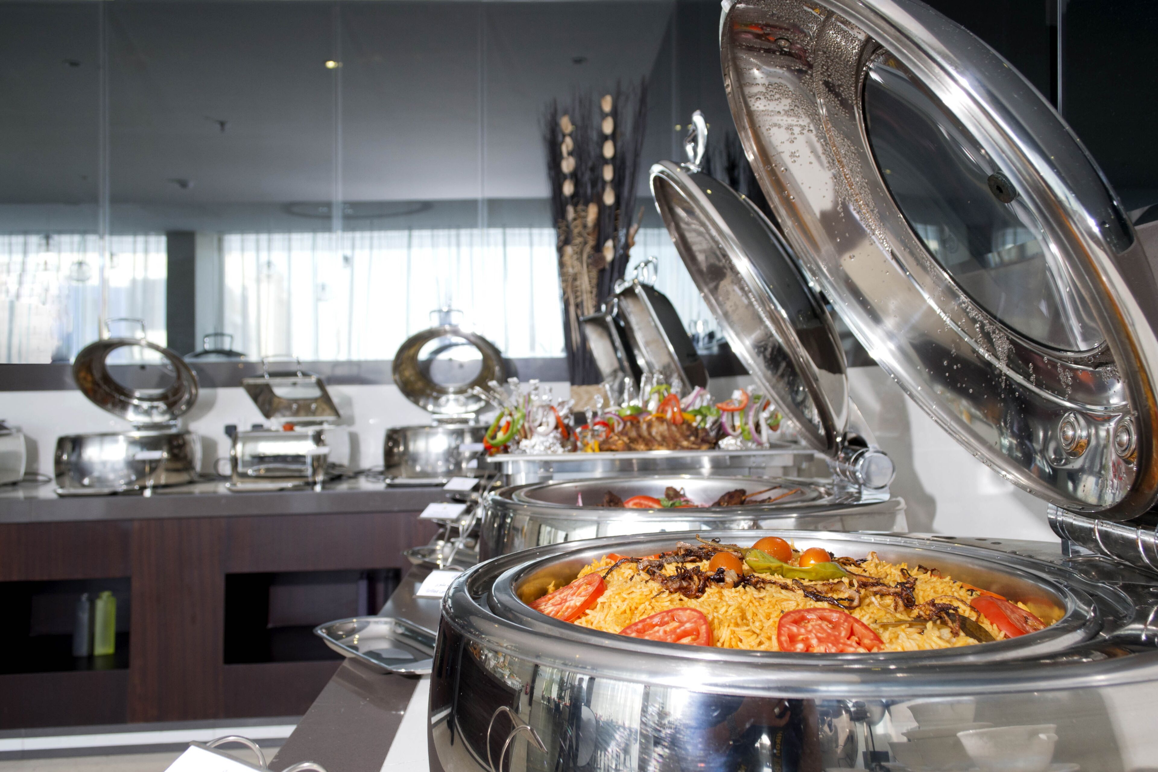 A buffet setup with multiple stainless steel serving dishes, some with lids open, revealing rice dishes garnished with vegetables.