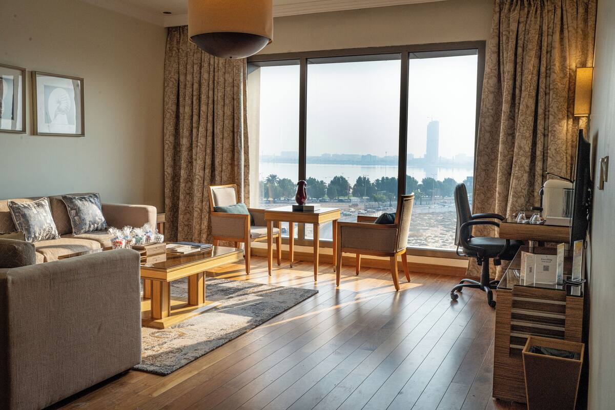 Ein modernes Wohnzimmer mit Sofa, Couchtisch und Sesseln, einem Schreibtisch mit Computer sowie einem großen Fenster mit Blick auf eine Stadtansicht.