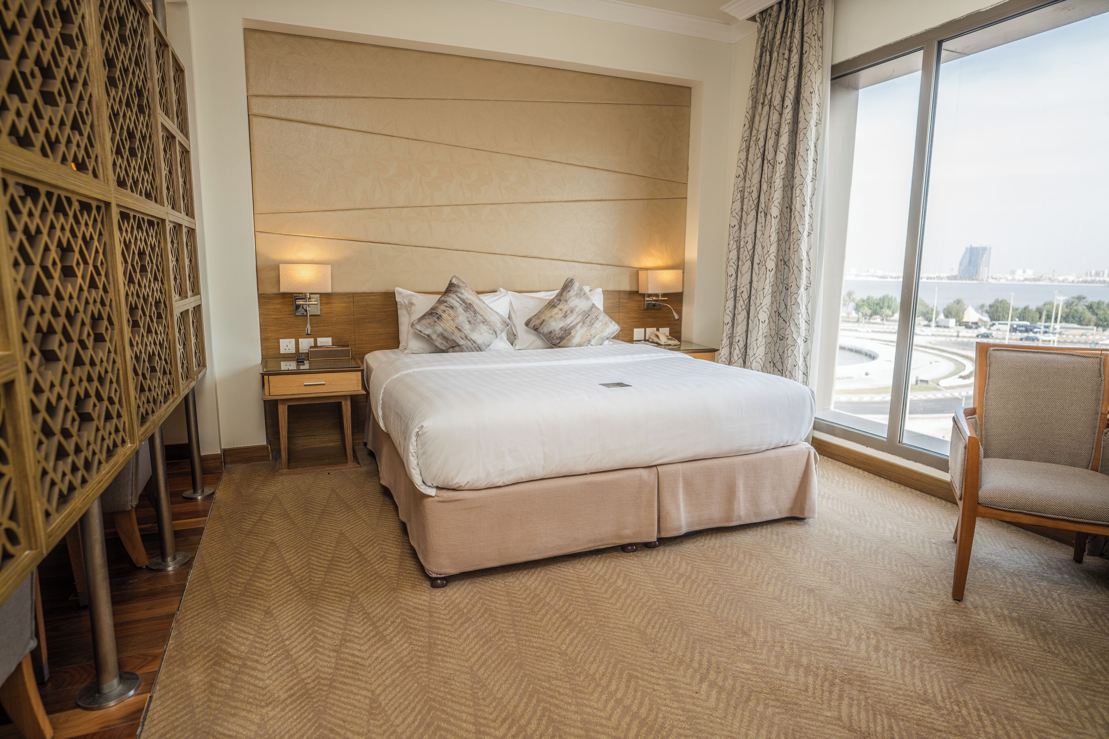 A hotel room with a large bed, a chair, a nightstand, and a view of a cityscape through a large window.