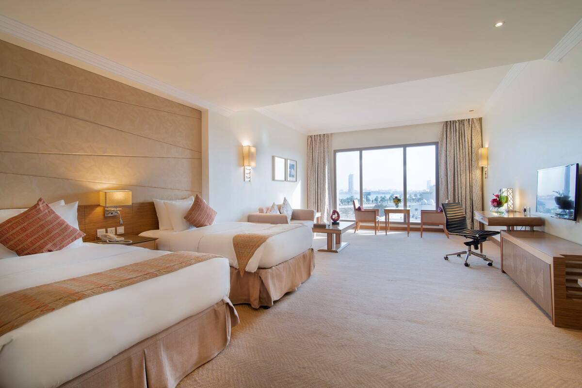 Ein Hotelzimmer mit zwei Betten, einem Schreibtisch, einem Stuhl und einem großen Fenster mit Blick auf die Stadt.