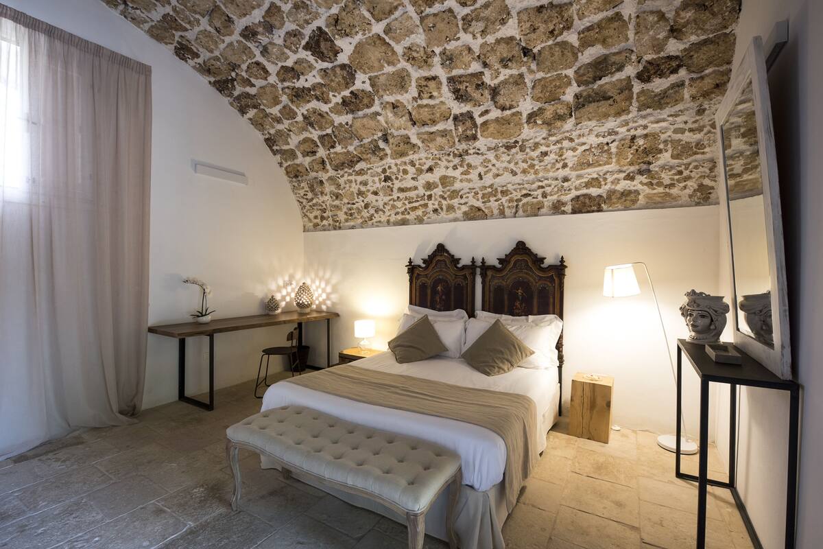 A bedroom with a stone ceiling, a large bed with a wooden headboard, a bench, bedside tables with lamps, and a small wooden stool.