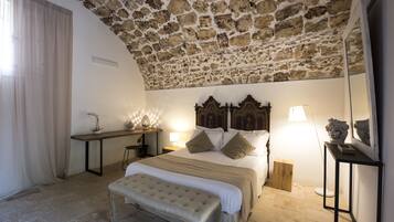 Una camera da letto con soffitto in pietra, un letto grande con testiera in legno, una panca, comodini con lampade e uno sgabello di legno.