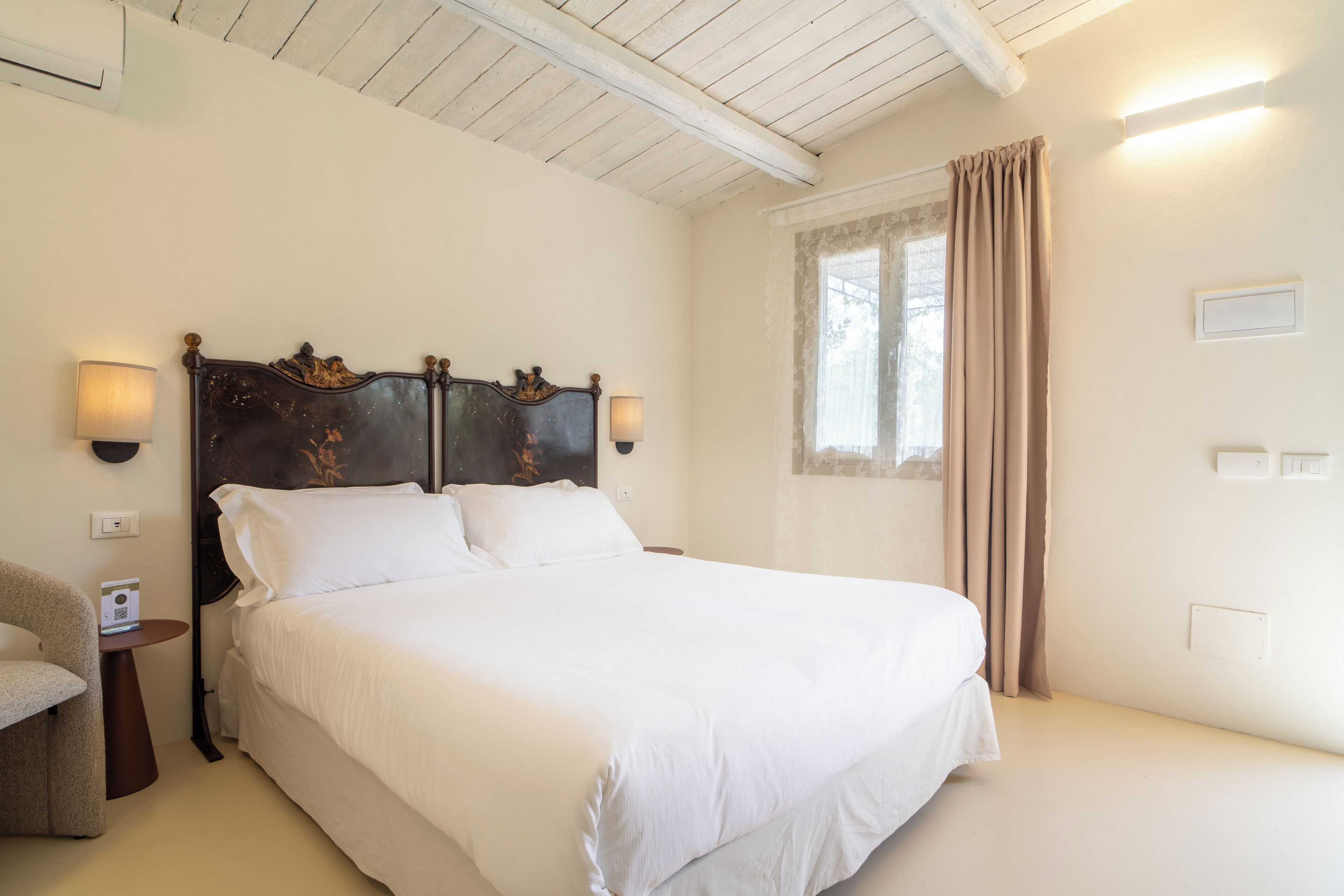 A bedroom with a large bed, two bedside lamps, a chair, a small table, and a window with curtains.