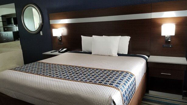 A hotel room with a large bed, a bedside table with a phone, a mirror, and a nightstand with a lamp. - HomeBridge Inn and Suites (Beaumont)