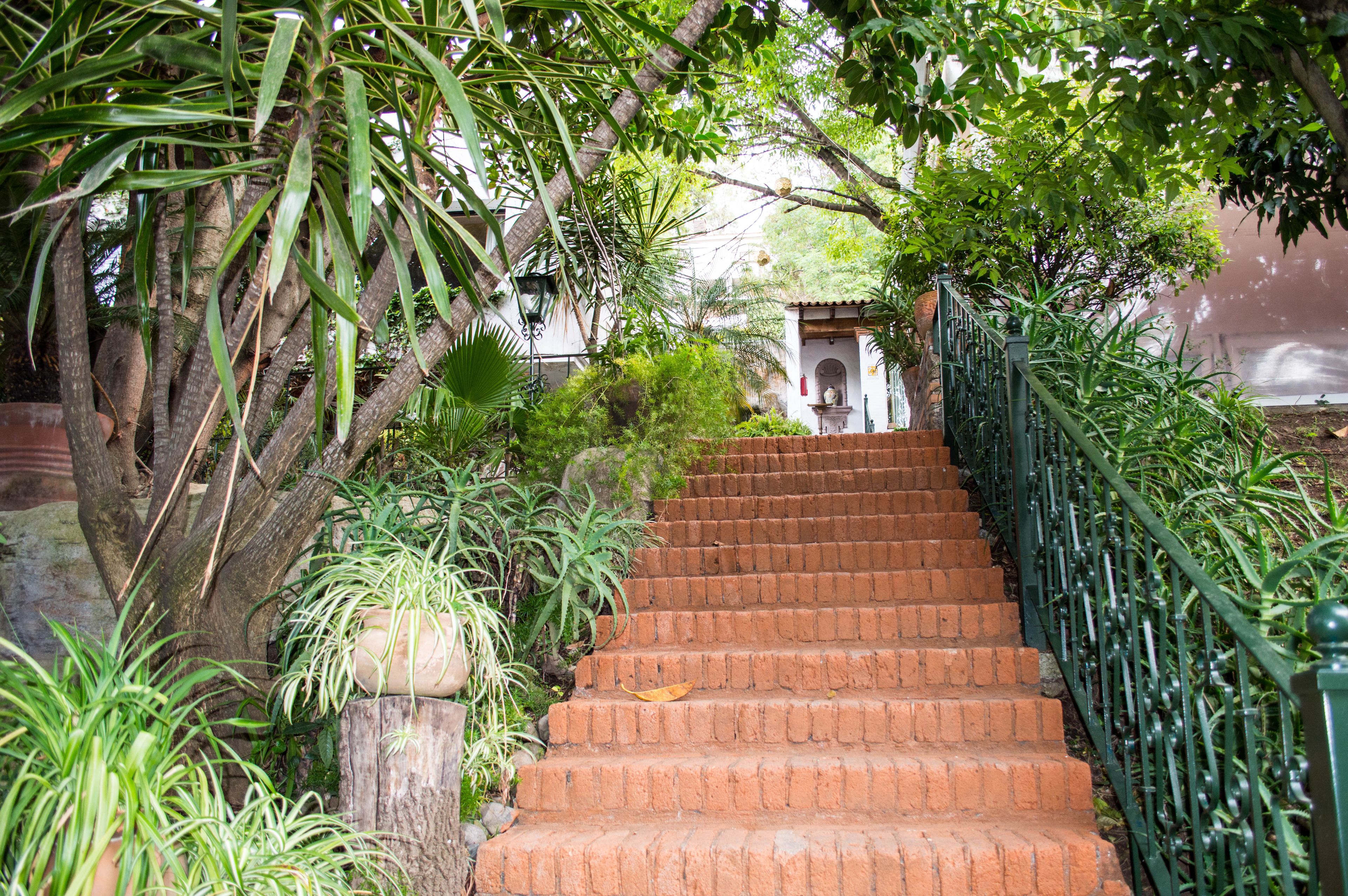 A brick staircase with a green metal railing, surrounded by lush greenery and a small white building in the background.