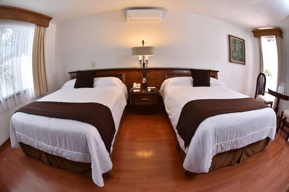 Une chambre d’hôtel avec deux lits, une table de chevet avec une lampe, un tableau au mur et une fenêtre avec des rideaux.