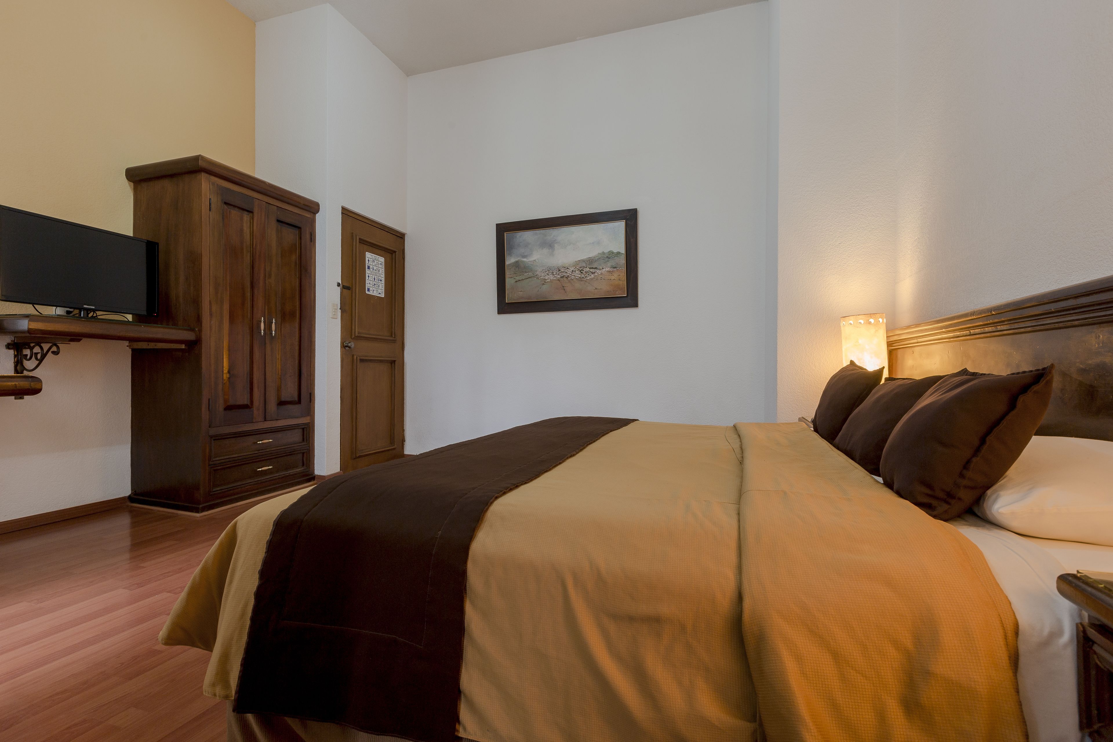 A hotel room with a bed, a television, a wardrobe, and a painting on the wall.