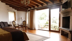 Un dormitorio amplio con una cama grande, suelo de madera, chimenea, un balcón con vistas a la vegetación y un comedor con sillas y una mesa.