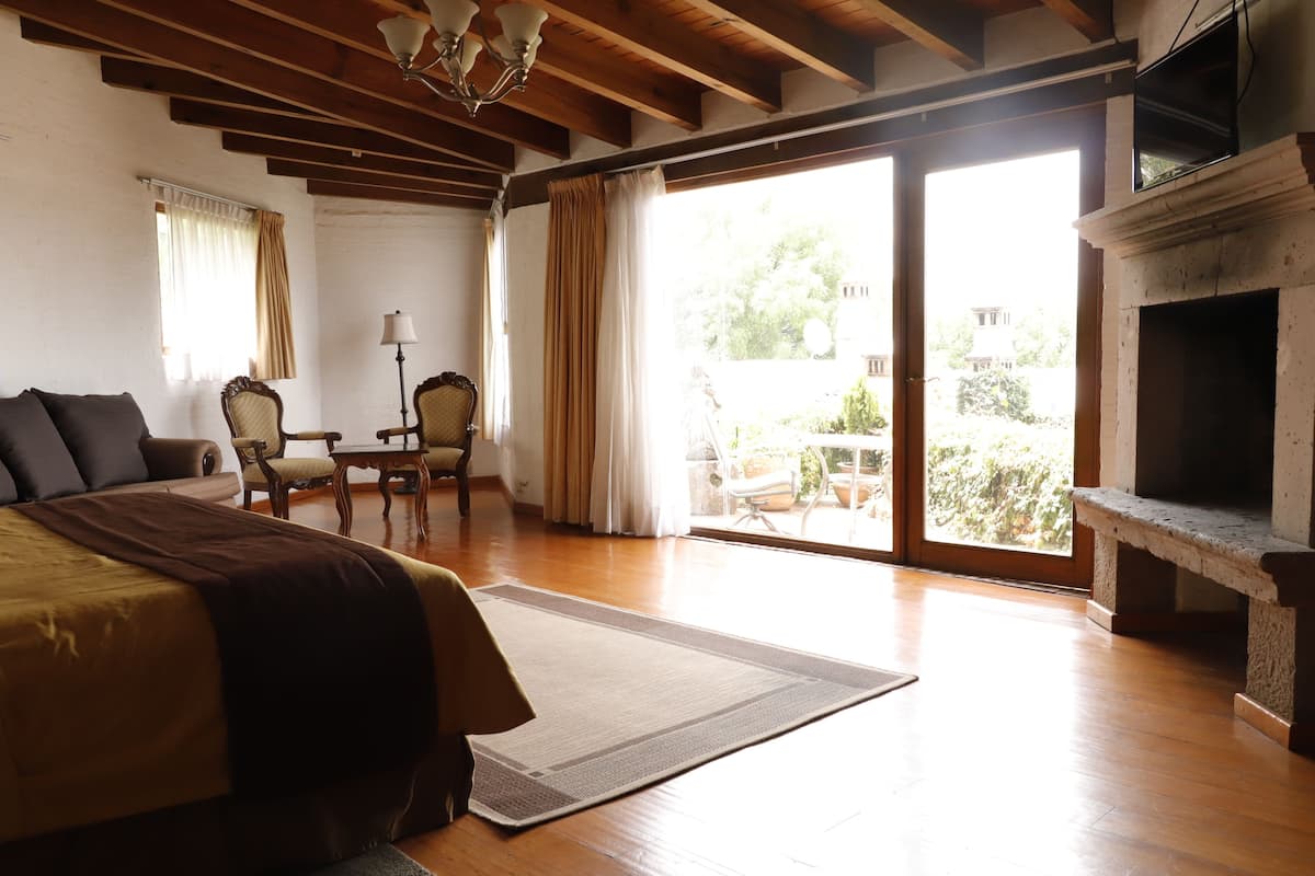 Une chambre spacieuse avec un grand lit, un plancher en bois, une cheminée, un balcon offrant une vue sur la verdure et un coin repas avec des chaises et une table.