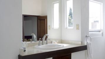 Un baño con lavamanos blanco, encimera oscura y espejo. Dos ventanas permiten la entrada de luz natural.