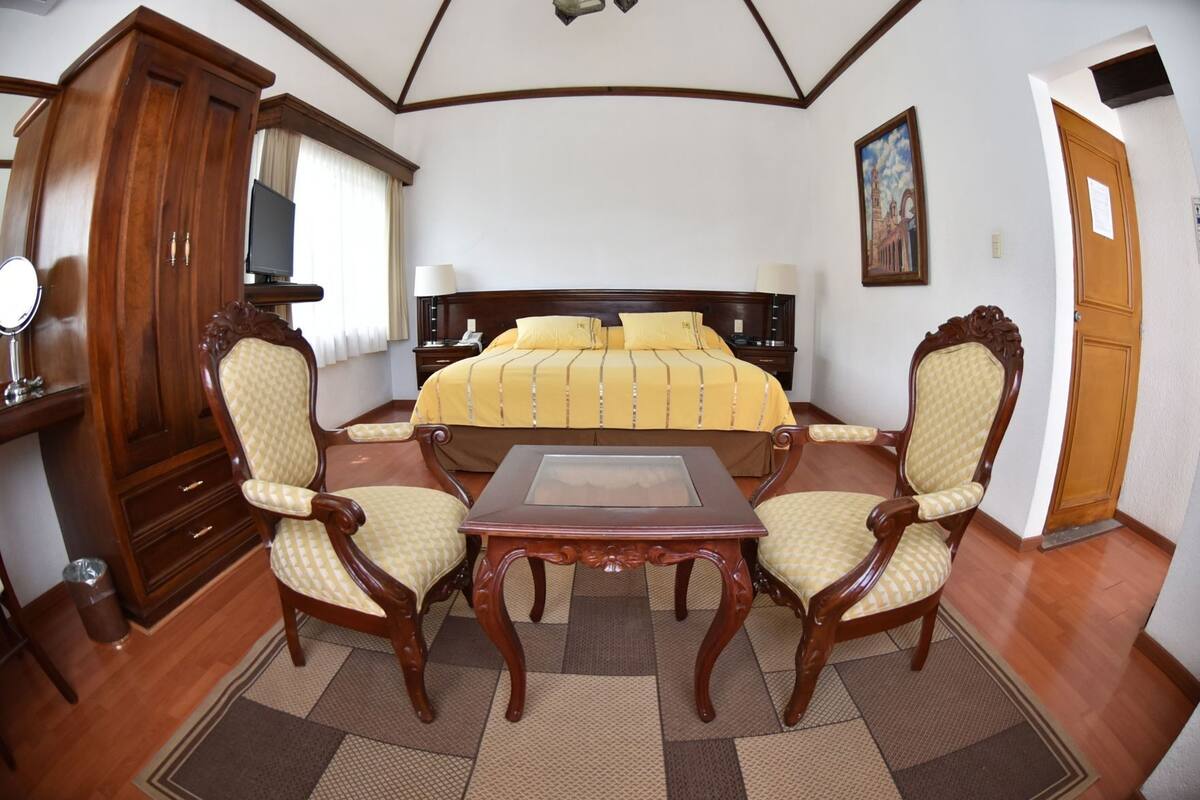 Une chambre d’hôtel avec un grand lit, deux fauteuils, une petite table, une armoire, un miroir et un tableau au mur.