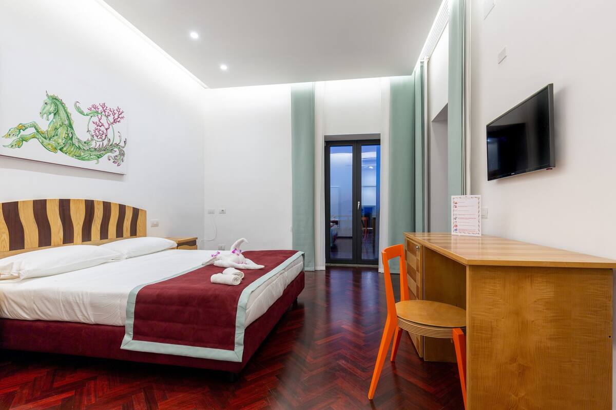 A hotel room with a bed, a desk, a chair, a TV, and a painting on the wall.
