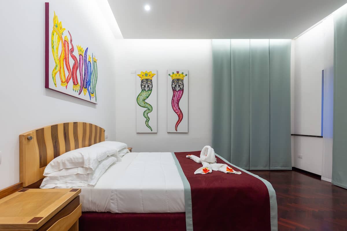 A hotel room with a wooden headboard, a bed with white linens, a red throw blanket, a wooden side table, and two wall-mounted artworks depicting stylized snakes with crowns.