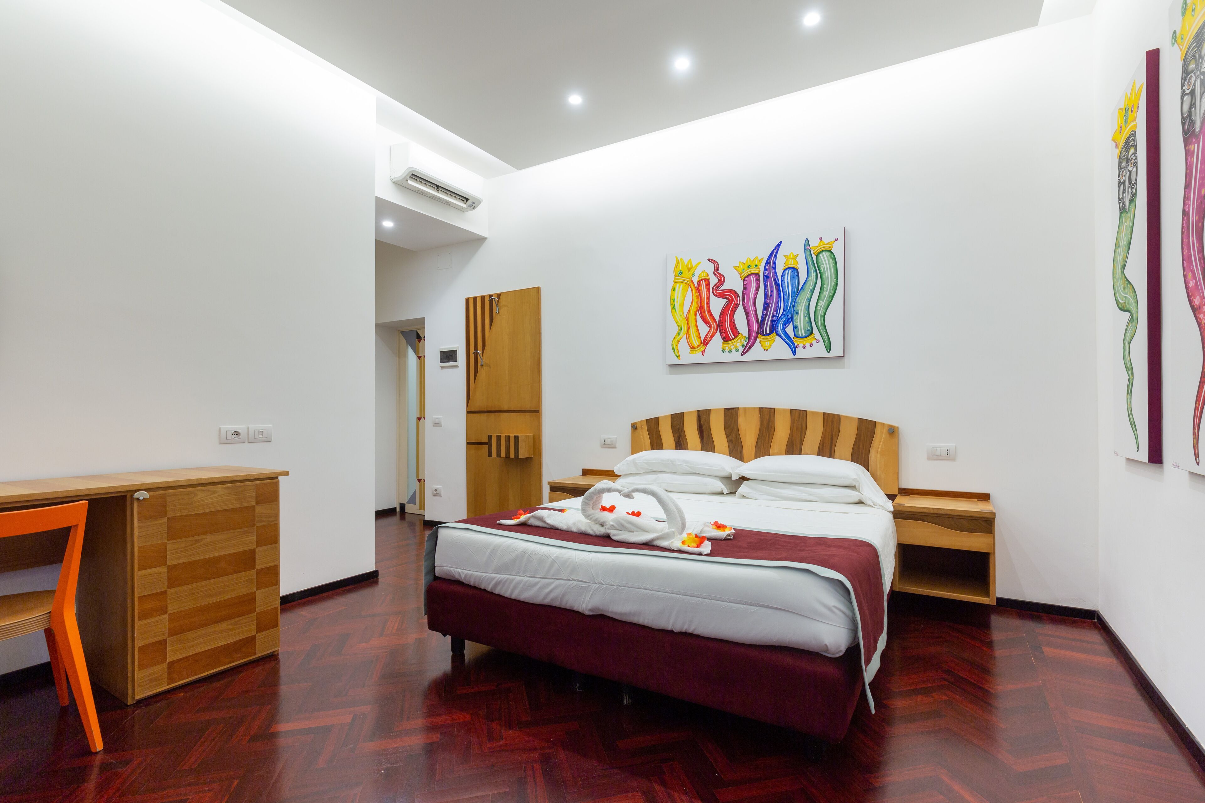 Une chambre d’hôtel moderne avec un bureau en bois, un lit recouvert d’une courtepointe rouge et un mur orné d’œuvres d’art colorées.