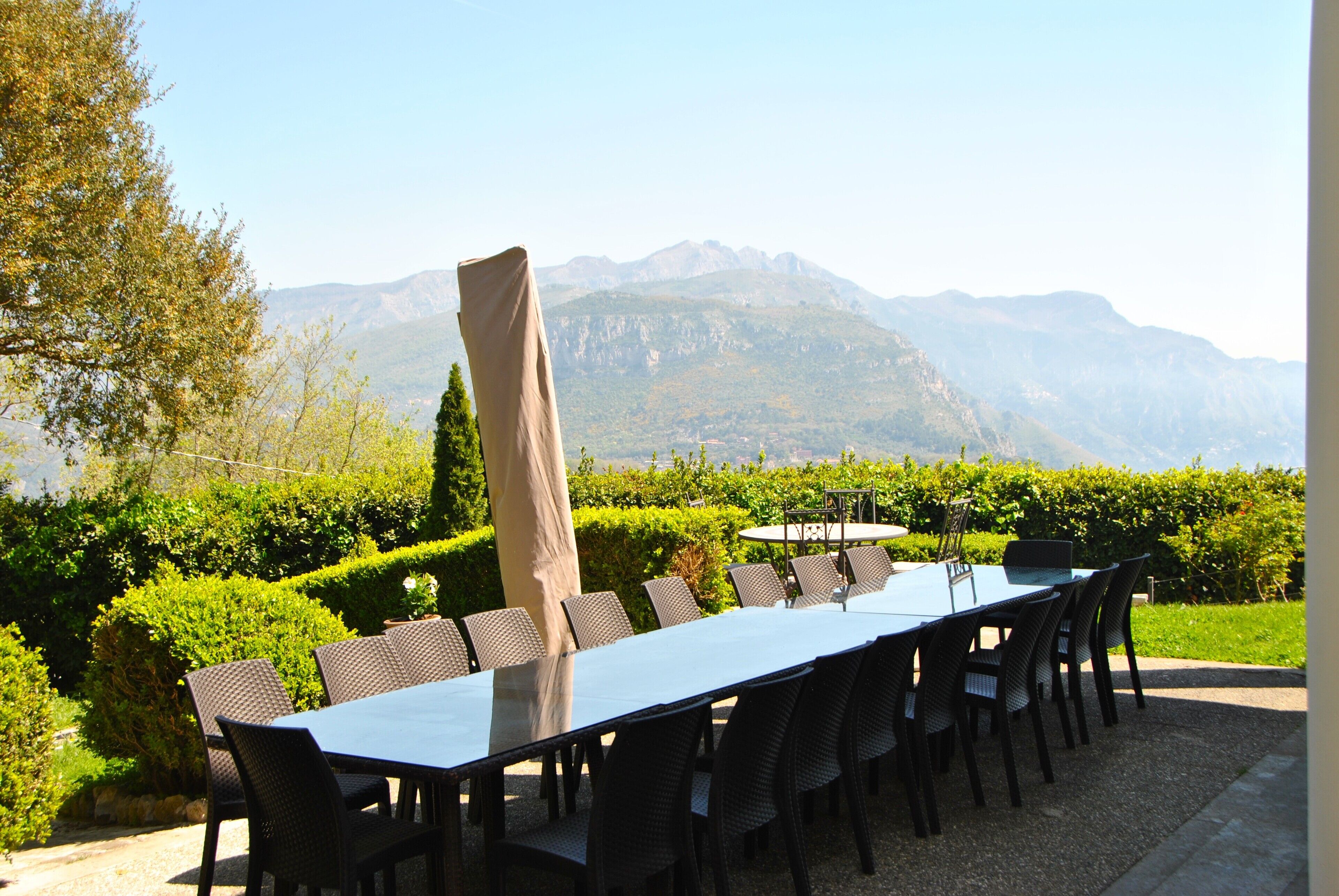An outdoor dining area with a long table and chairs, surrounded by greenery and a mountain view.