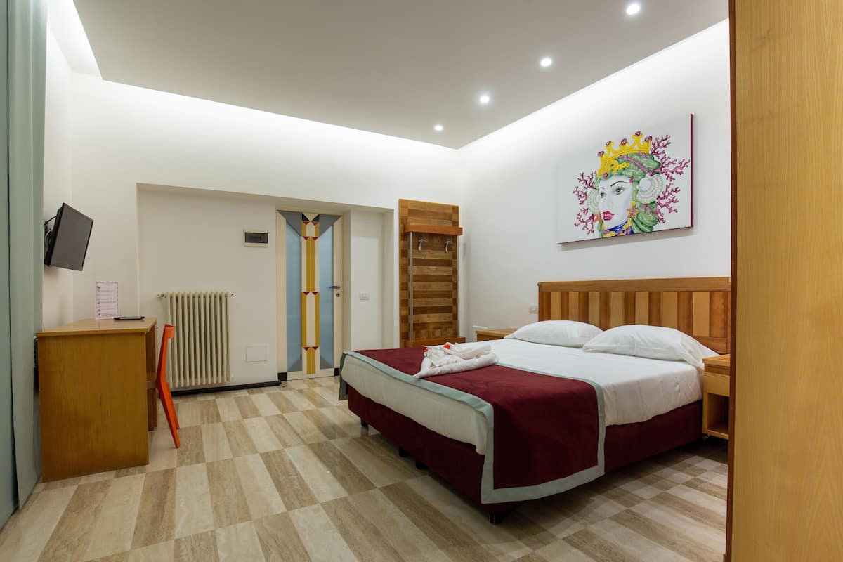 A hotel room with a bed, a wooden headboard, a nightstand, a desk, a chair, a radiator, and a painting on the wall.