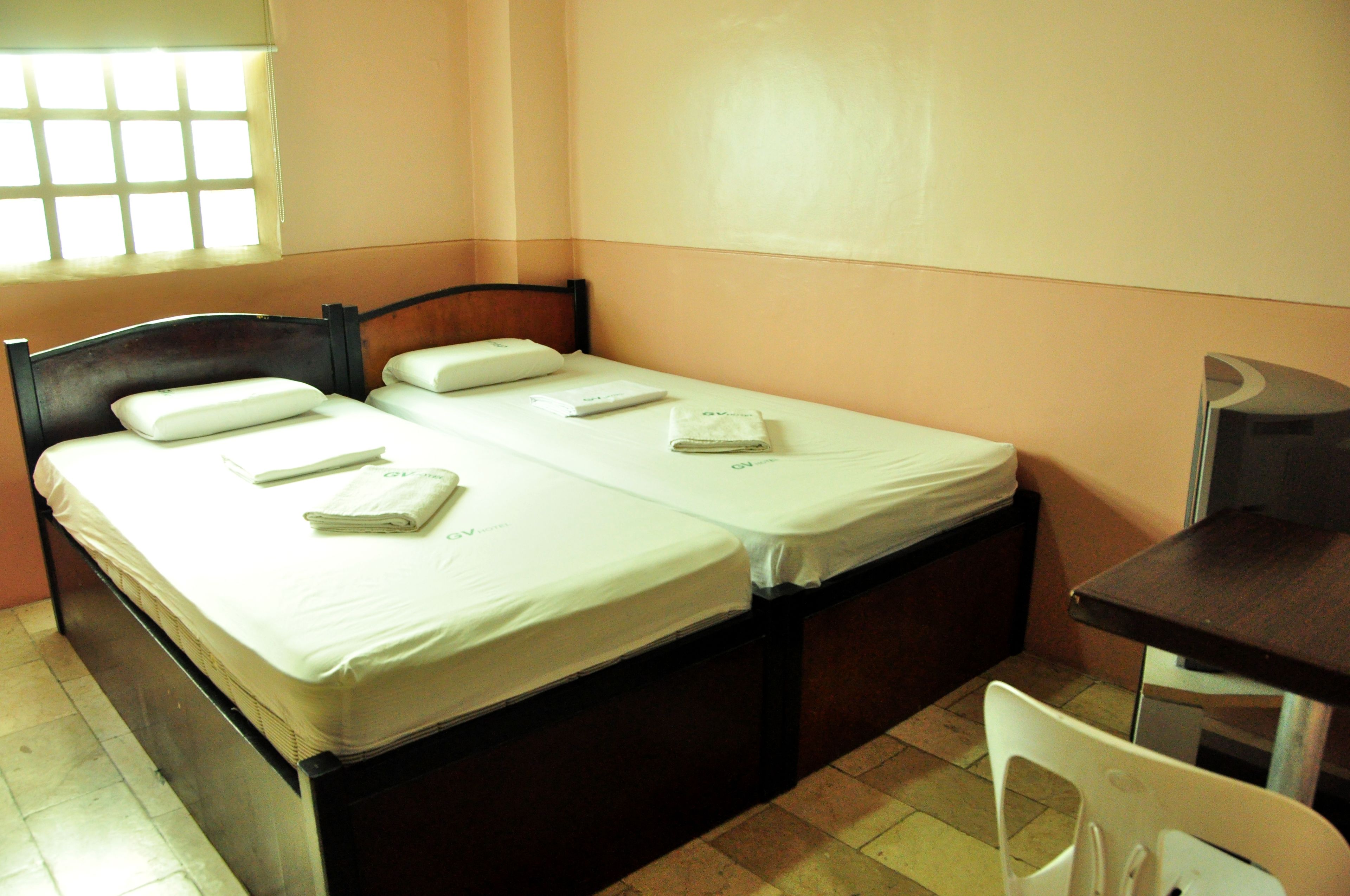 Una cama individual con cabecera de madera, una mesita pequeña y una silla en una habitación de distribución sencilla y una ventana con diseño de cuadrícula.
