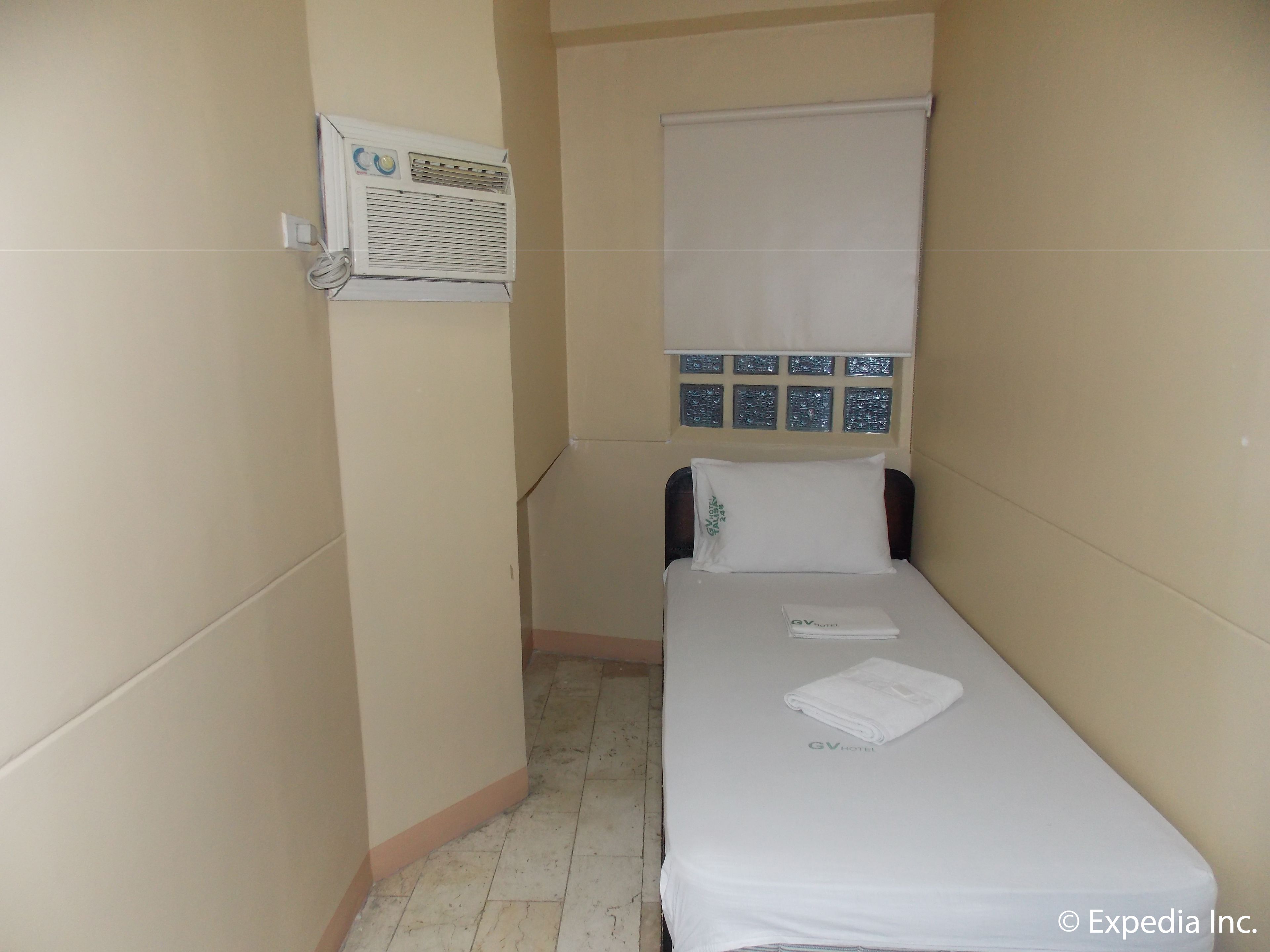 Una habitación de hotel pequeña y estrecha con una cama individual, una ventana con persianas, un aire acondicionado de pared y un salpicadero de azulejos sobre la cama.