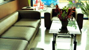 A hotel lobby with a sofa, armchairs, a coffee table with a flower vase, a television mounted on the wall, and a view of the street outside. - GV Hotels Talisay City (Talisay)