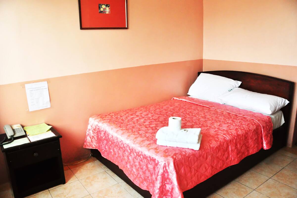 A neatly made bed with a red bedspread, a white pillow, and a white towel on the side. There is a small black nightstand with a phone and papers on it.
