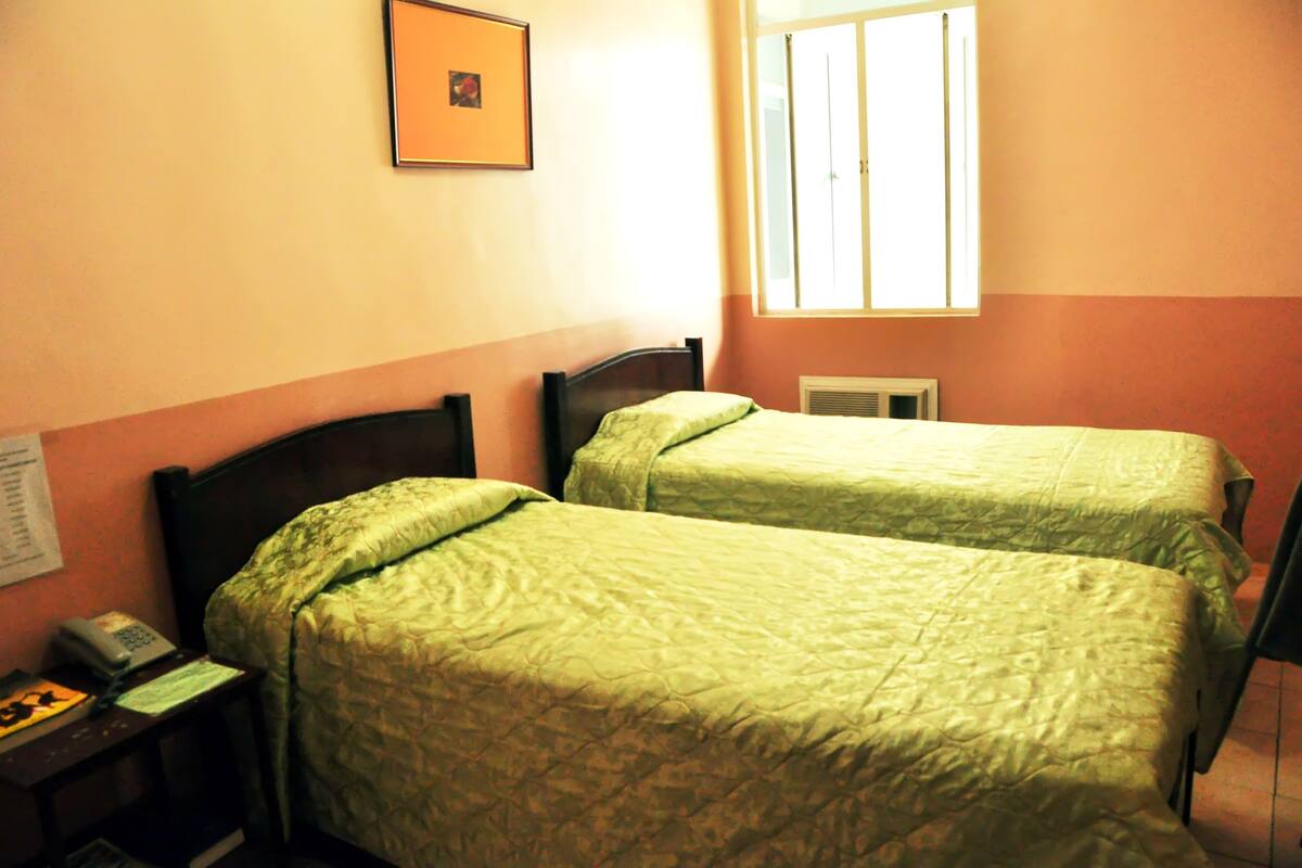 A room with two single beds, a bedside table with a telephone, and a window with blinds.
