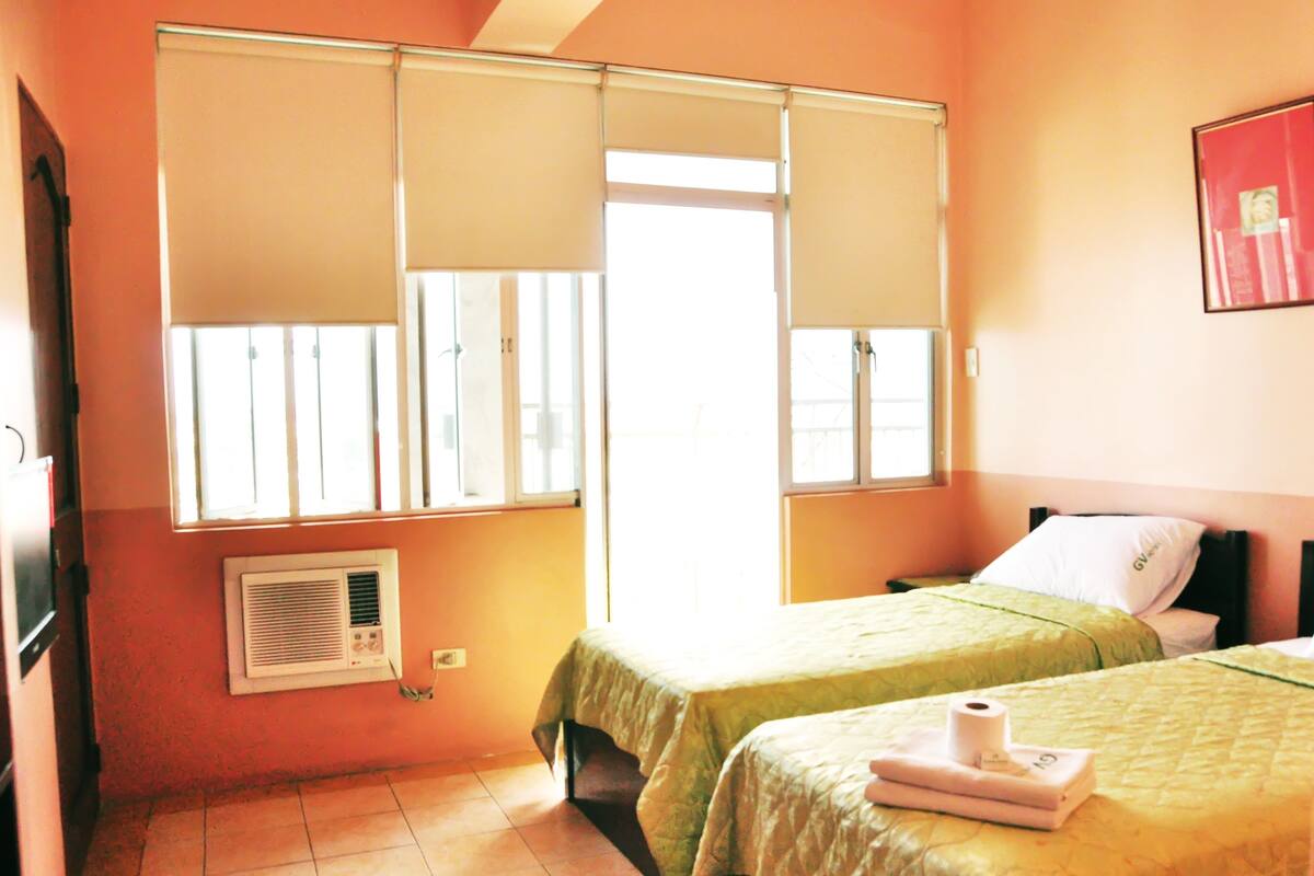 A room with two beds, a window with blinds, a wall-mounted air conditioner, and a framed picture on the wall.