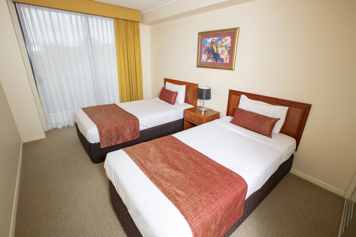 Habitación de hotel con dos camas, una mesita de noche, una lámpara, un cuadro en la pared y una ventana con cortinas.