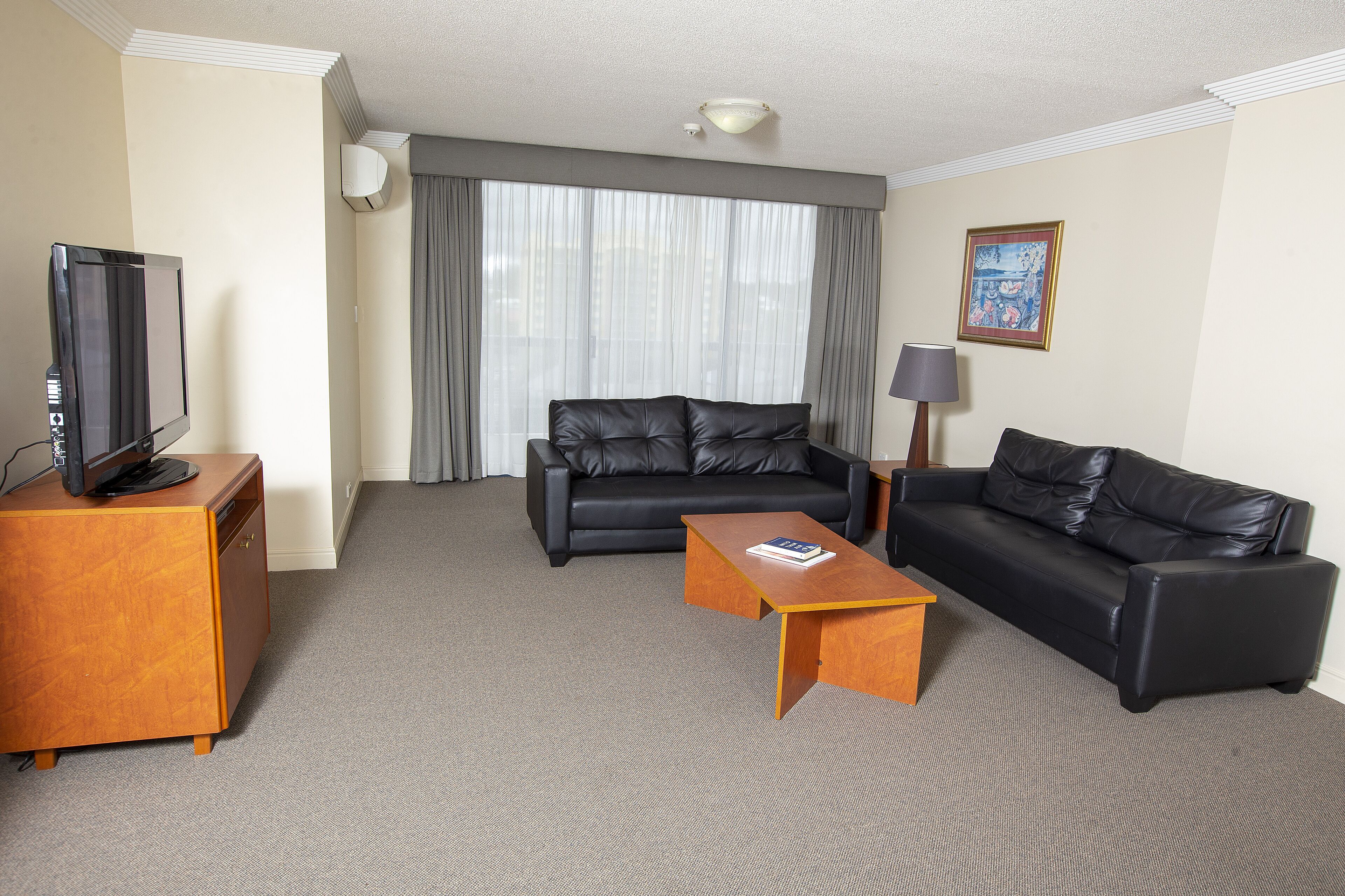 A hotel room with a black leather sofa, a wooden coffee table, a TV on a wooden cabinet, and a large window with curtains.