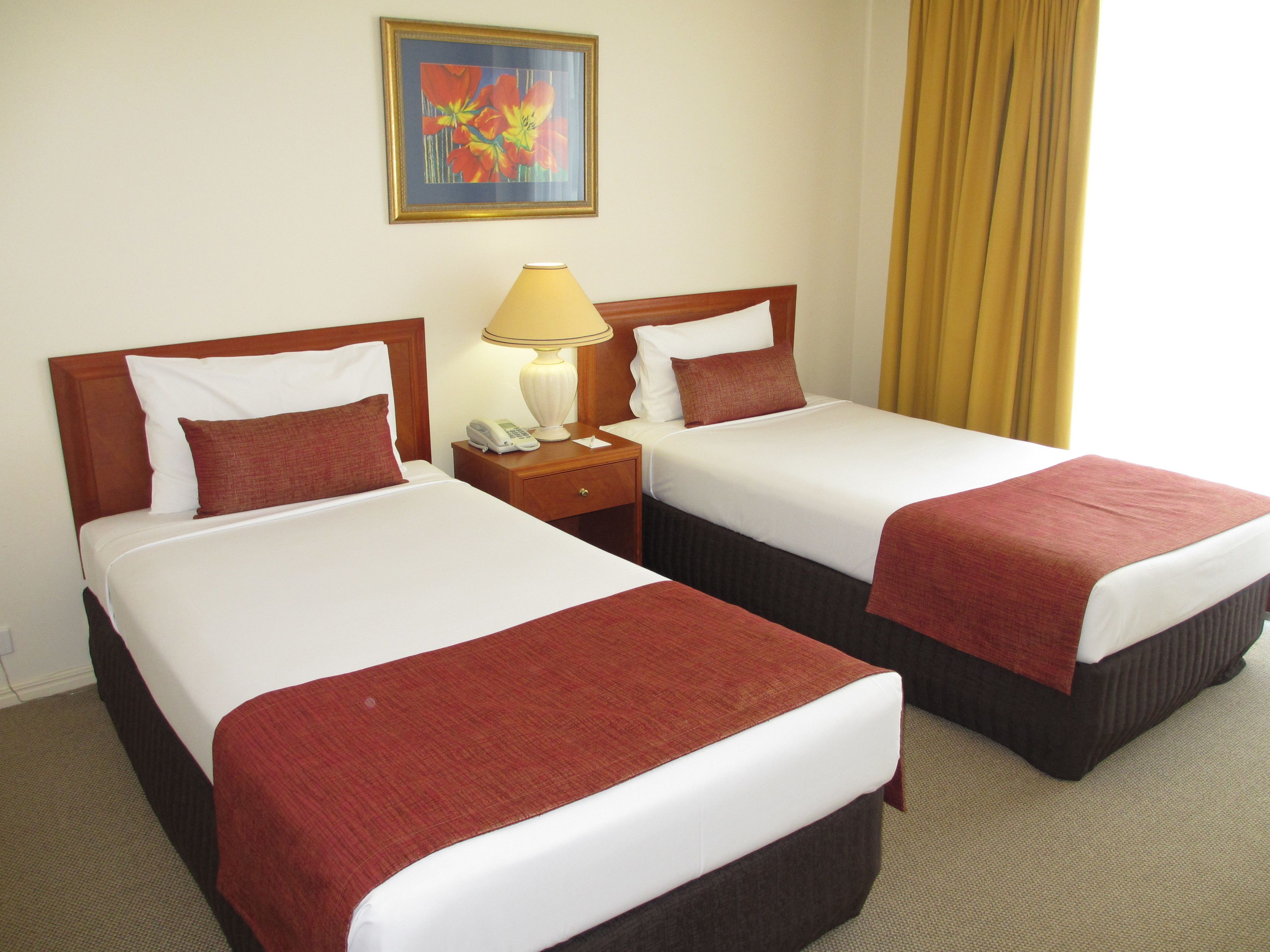 Habitación de hotel con dos camas, cada una con una manta roja, sábanas blancas y cabeceros de madera. Entre las camas hay una mesita de noche con una lámpara y un teléfono. Un cuadro cuelga de la pared sobre las camas.