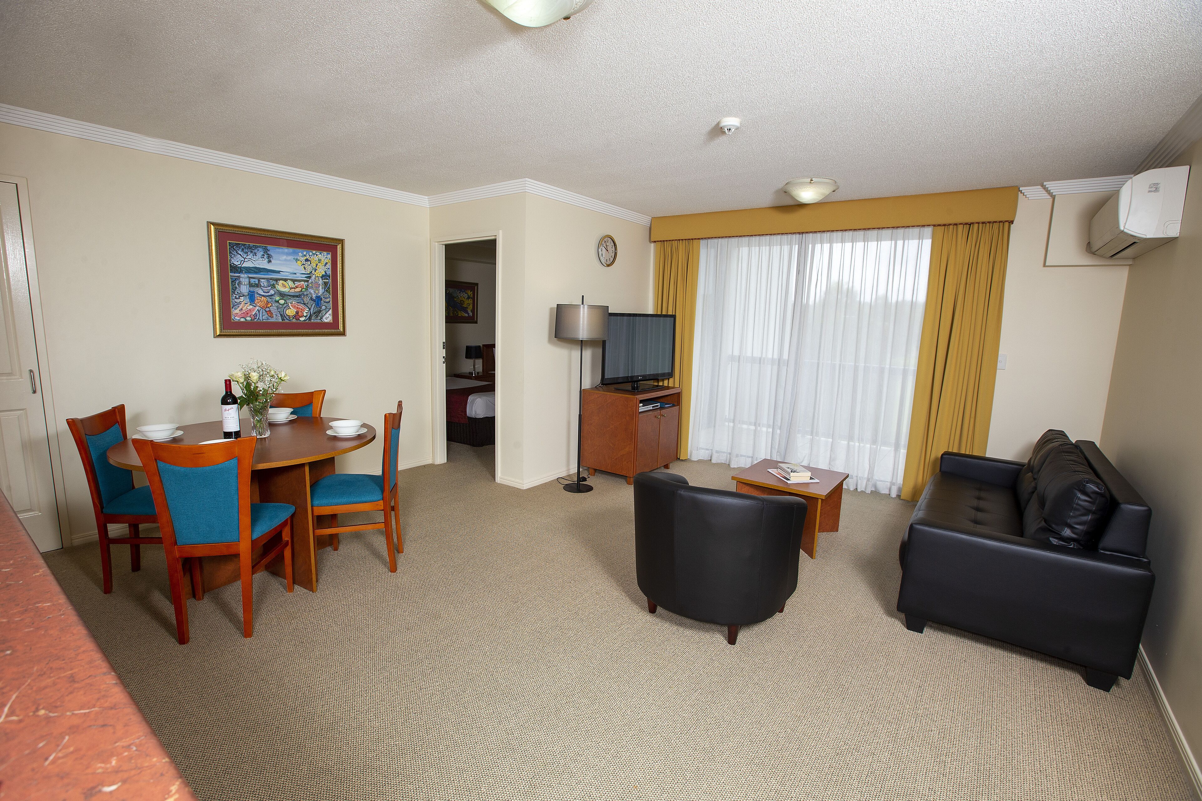 A hotel room with a dining area, a living room with a black leather sofa, a TV, and a painting on the wall.