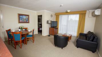 A hotel room with a dining area, a living room with a black leather sofa, a TV, and a painting on the wall.