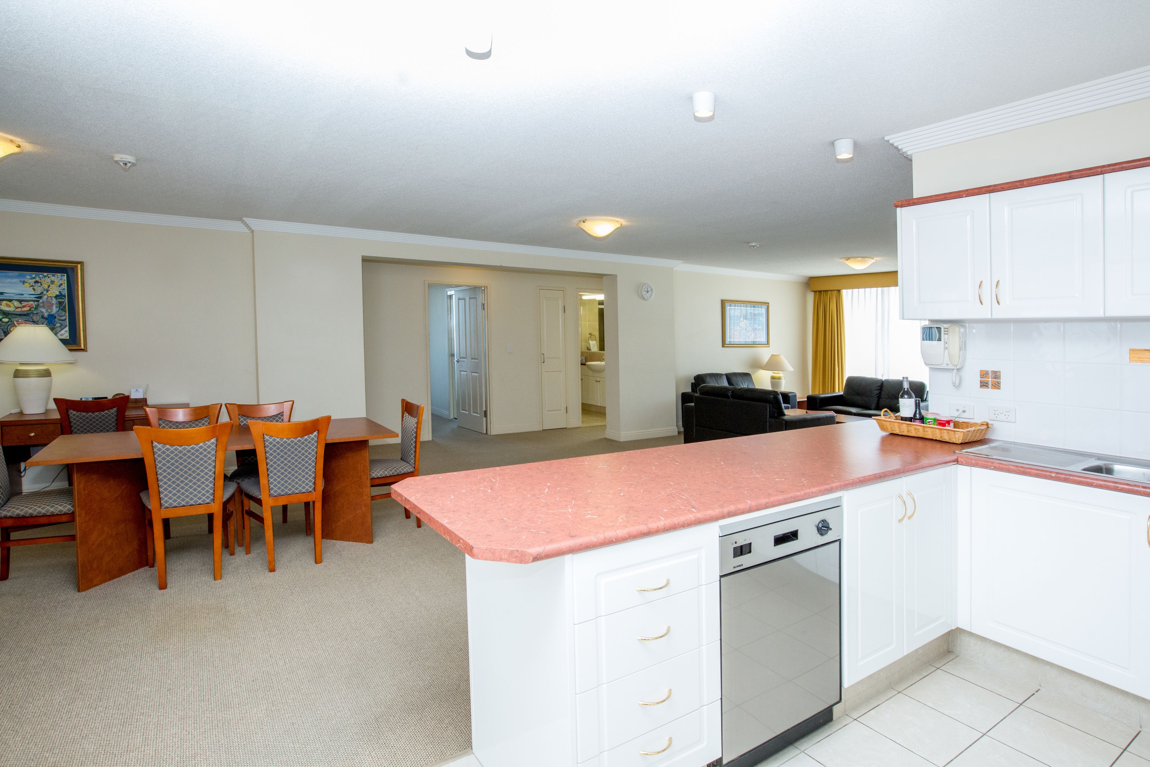 A modern kitchen with white cabinets, a granite countertop, and a dishwasher. There is a dining area with wooden chairs and a table, and a living room with a sofa and a coffee table.