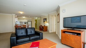 A living room with a black leather sofa, a wooden coffee table with a red book, a TV on a wooden stand, and a kitchen in the background. - Springwood Tower Apartment Hotel (Springwood)
