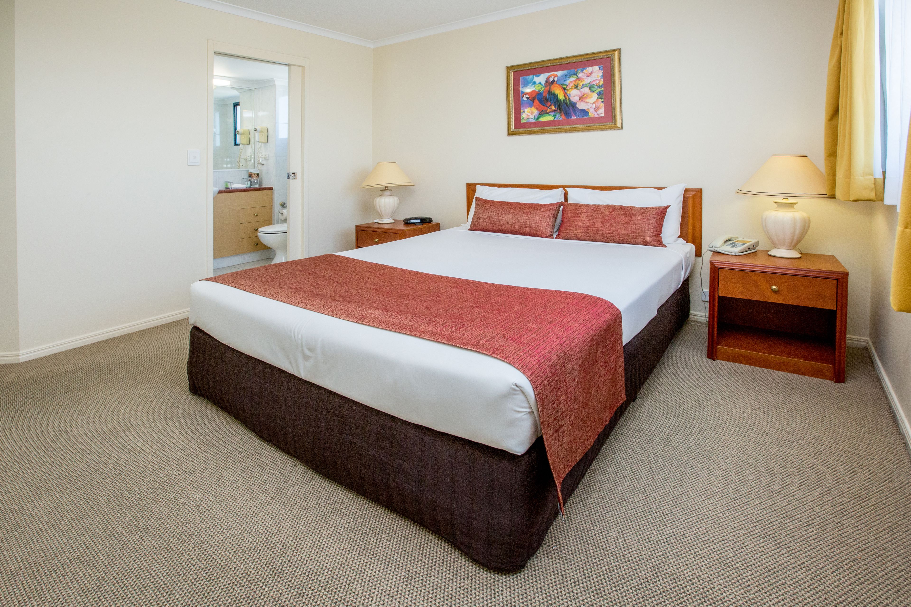 A hotel room with a large bed, two bedside tables, a nightstand, a painting on the wall, and a bathroom visible through an open door.