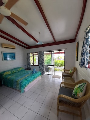 A room with a bed, two wicker chairs, a ceiling fan, and a view through a glass door. - Reefcomber Sunset Beach (Rarotonga)