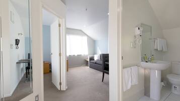 Una habitación de hotel moderna con un espejo grande, un lavabo blanco y un inodoro. Hay un sofá gris y una pequeña mesa en la esquina.