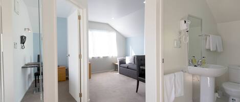 A modern hotel room with a large mirror, a white sink, and a toilet. There is a grey sofa and a small table in the corner.