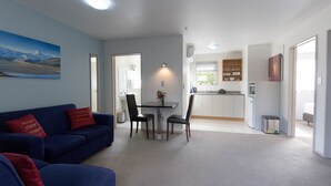 Een moderne woonkamer met een blauwe bank en een eethoek met tafel en stoelen. In de achtergrond is een keuken met witte kasten en een raam zichtbaar.
