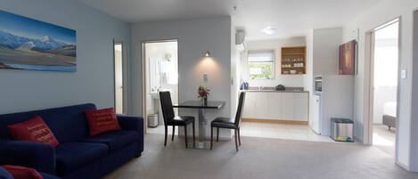 Un salon moderne avec un canapé bleu et un coin repas comprenant une table et des chaises. On aperçoit, au fond, une cuisine avec des meubles blancs et une fenêtre.