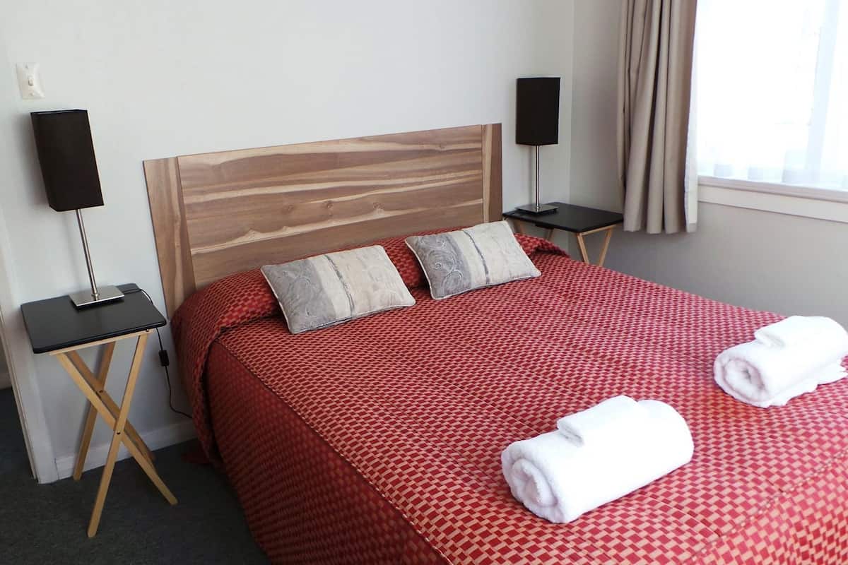 A hotel room with a wooden headboard, two bedside tables with lamps, a red patterned bedspread, and two rolled towels on the bed.