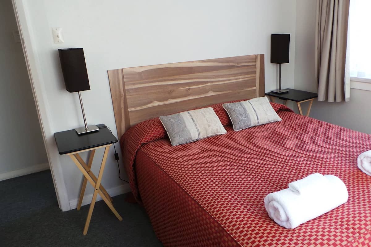 A hotel room with a wooden headboard, two bedside tables, a red patterned bedspread, two pillows, and two rolled towels.