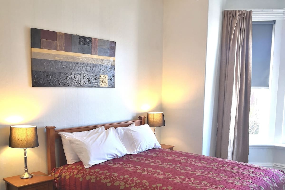 A hotel room with a large bed, two bedside lamps, a chandelier, a window with curtains, and a piece of abstract art on the wall.