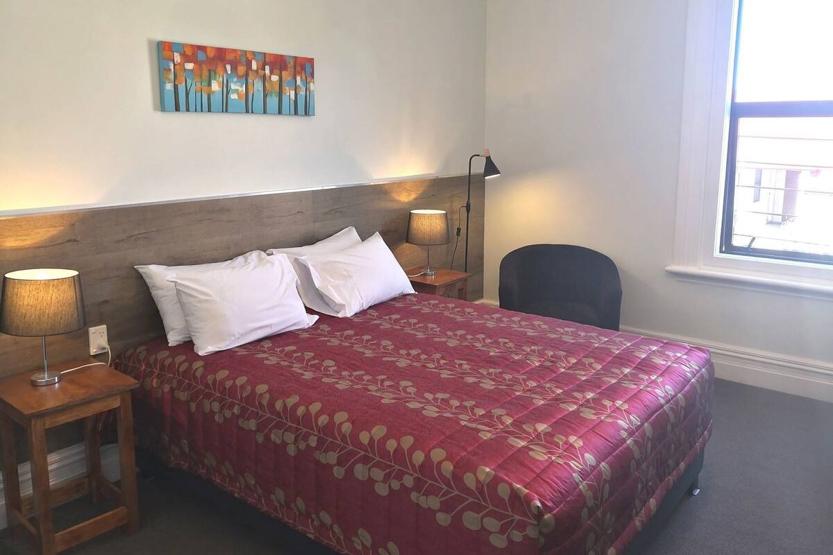 A hotel room with a bed, two bedside lamps, a chair, a window, and a wall art piece.