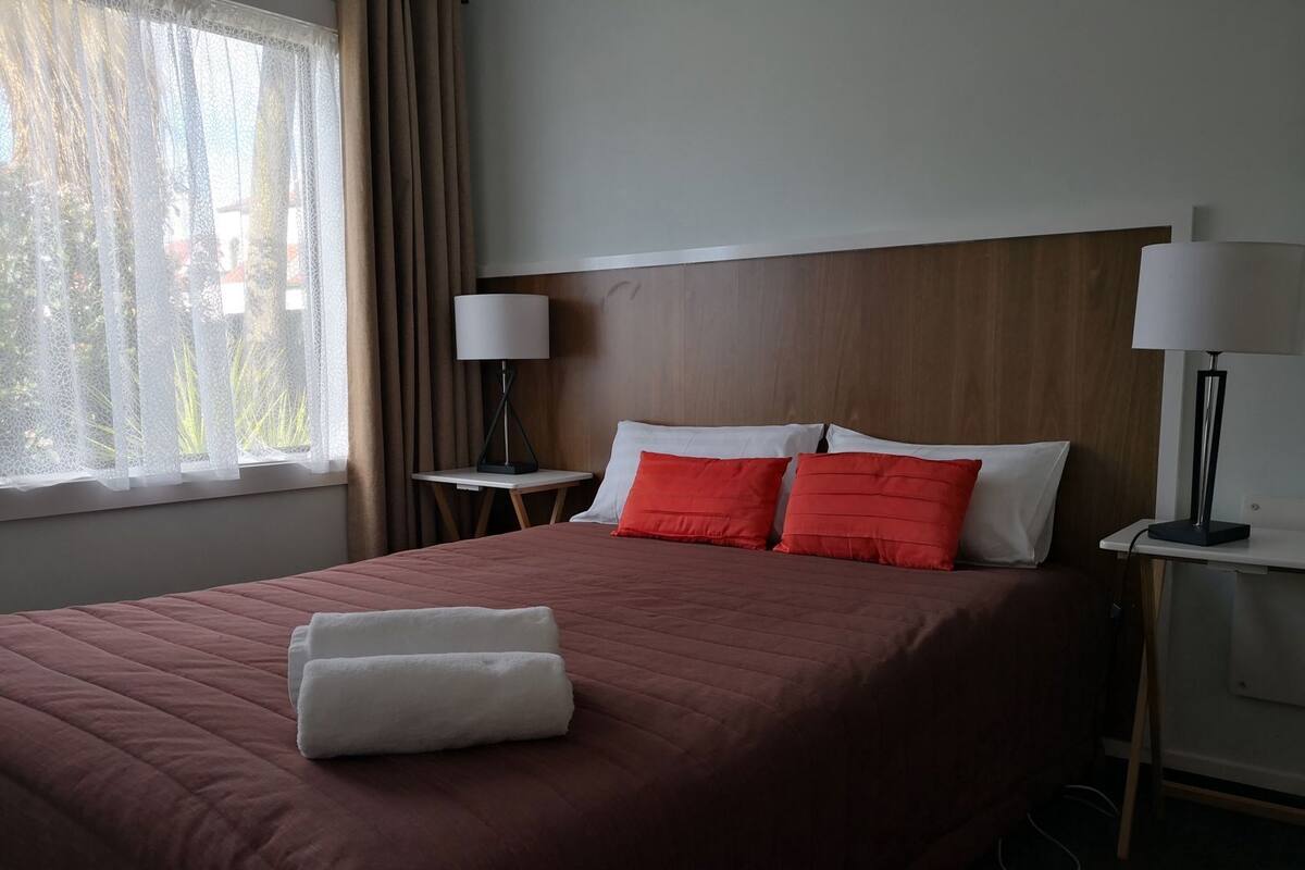 A hotel room with a large bed, two bedside tables with lamps, a window with curtains, and a wooden headboard.